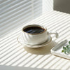 A white ceramic coffee cup with a gold rim sits on a matching saucer filled with black coffee, placed on a sunlit white table beside a small white dish holding green eucalyptus leaves. Parallel window blinds cast diagonal shadows across the scene.