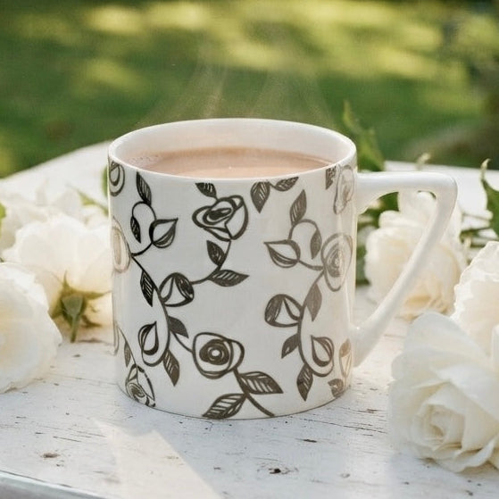 A white ceramic mug with a silver rose floral pattern sits on a white wooden table outdoors, surrounded by white roses, with a blurred green garden background.