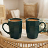 Two dark green ceramic mugs filled with coffee on a woven tray in a warm, softly lit room.