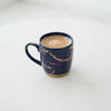 A dark blue ceramic mug with gold marbled veins and a gold-toned base, filled with a light brown coffee drink, set against a clean white background.