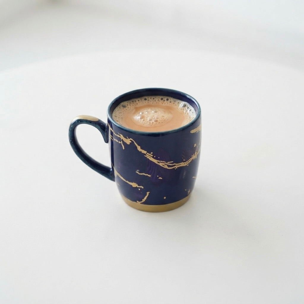 A dark blue ceramic mug with gold marbled veins and a gold-toned base, filled with a light brown coffee drink, set against a clean white background.