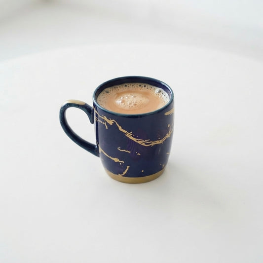 A dark blue ceramic mug with gold marbled veins and a gold-toned base, filled with a light brown coffee drink, set against a clean white background.