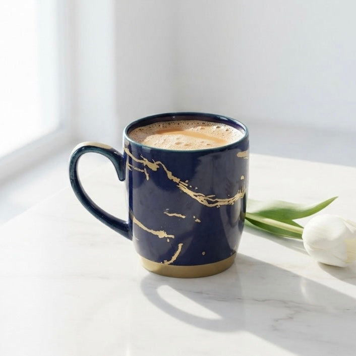 A navy blue ceramic mug with gold marble veining sits on a white marble table next to a white tulip, with a cup of coffee inside the mug.