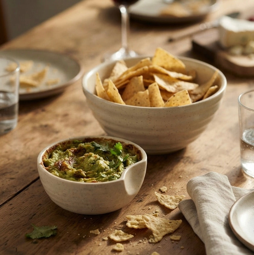 A rustic dining table setting featuring a small white ceramic bowl of guacamole and a larger bowl of tortilla chips, surrounded by plates, glasses of water, and a wine glass in a warm, natural light.