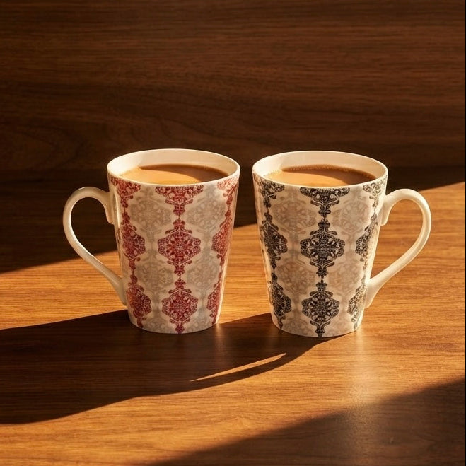 Two patterned ceramic mugs filled with coffee sit on a wooden table in warm sunlight, casting a long shadow. The mug on the left has a red and beige damask-style pattern, and the mug on the right has a dark gray and beige pattern.