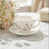 A white ceramic teacup and saucer set with a delicate taupe floral pattern, styled on a white marble table next to a pink rose bouquet.