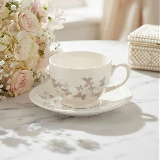 A white ceramic teacup and saucer set with a delicate taupe floral pattern, styled on a white marble table next to a pink rose bouquet.