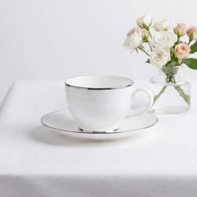 A white ceramic teacup and saucer with a thin metallic gold rim, styled on a white tablecloth next to a small glass vase of white and blush pink roses.