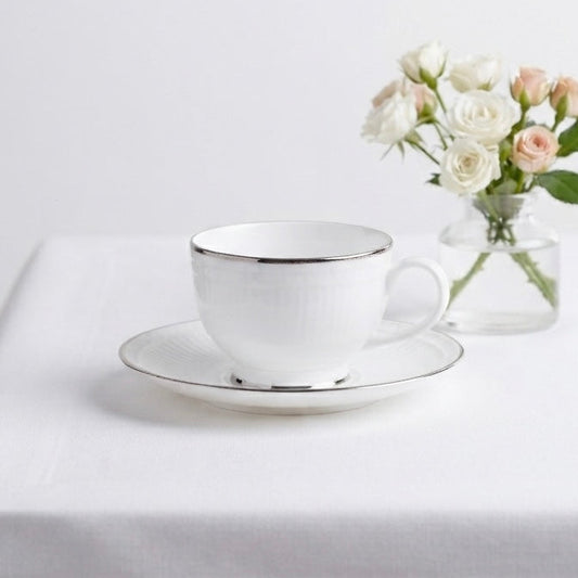 A white ceramic teacup and saucer with a thin metallic gold rim, styled on a white tablecloth next to a small glass vase of white and blush pink roses.