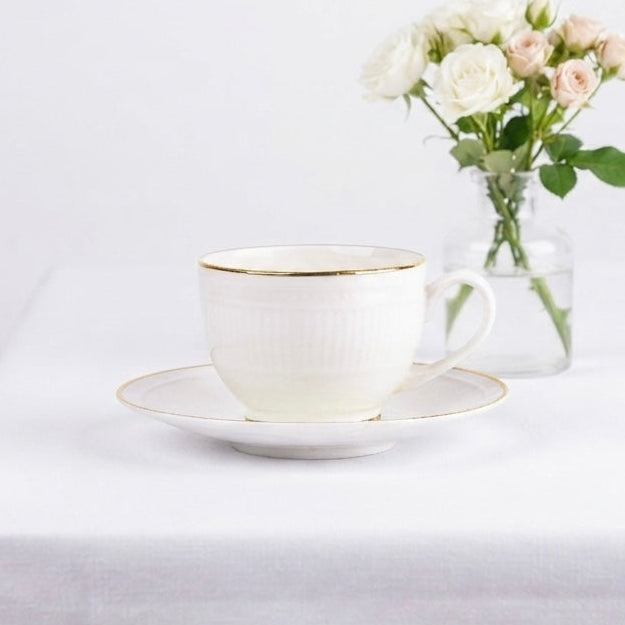 A white ceramic teacup and saucer with delicate gold trim, styled on a white tablecloth next to a small glass vase of white and blush pink roses against a white background.