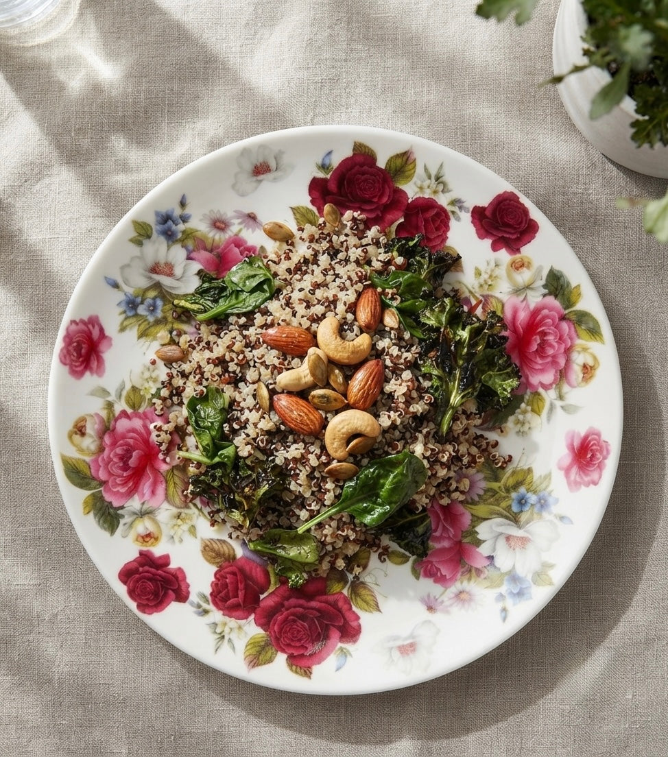 A floral ceramic plate filled with cooked quinoa, kale, almonds, and cashews, photographed from above on a beige linen surface with soft natural shadows.
