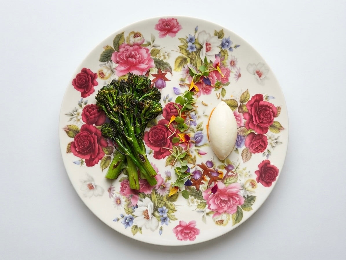 A round white ceramic plate with a vintage-style pink and green floral rose pattern, featuring a cooked broccoli stalk and a hard-boiled egg on top, photographed from a top-down view against a clean white background.