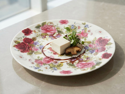 A round white ceramic plate with a pink and red rose floral pattern, styled with a small white cube, green microgreens, and a mushroom on a marble countertop.