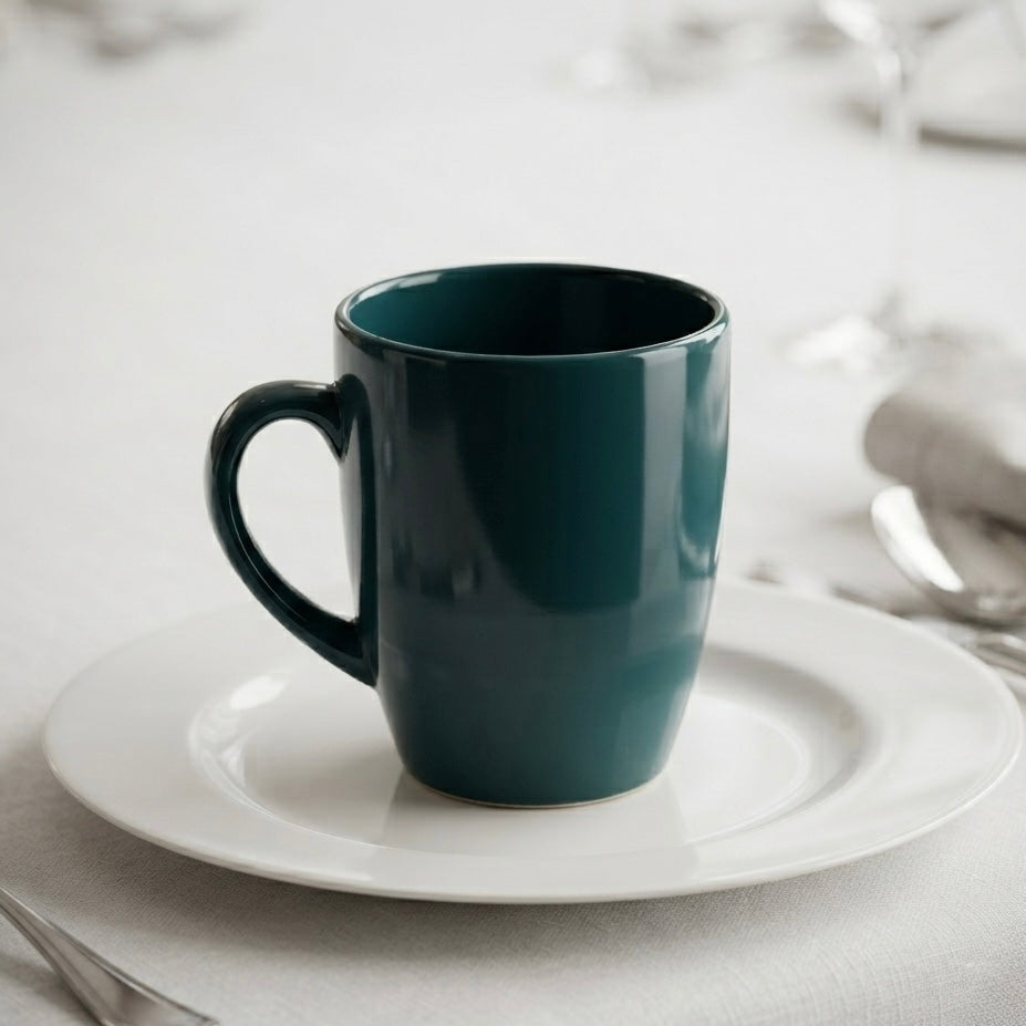A dark teal ceramic coffee mug, set on a white tablecloth with silver cutlery and blurred glassware in the background.