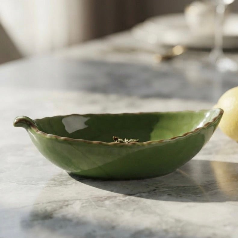 A green ceramic bowl with a scalloped rim and small side handles, styled on a marble table next to a lemon with a blurred dining background.