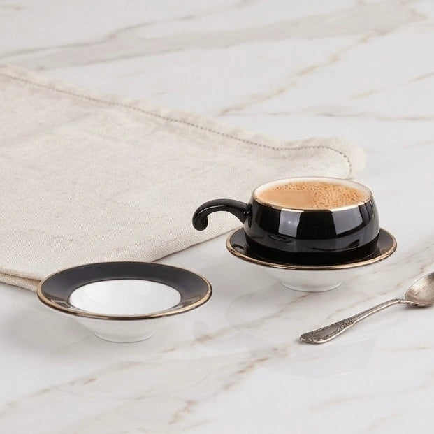 A small black coffee cup with a matching saucer, a small bowl, and a spoon arranged on a white marble table next to a beige cloth.