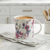 A white ceramic mug with a pink and purple floral pattern sits on a folded white napkin on a marble countertop in a bright kitchen. A small white jar with wooden spoons and a yellow lemon are nearby.