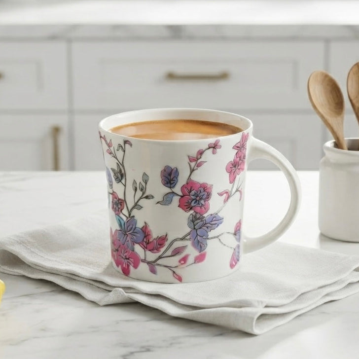 A white ceramic mug with a pink and purple floral pattern sits on a folded white napkin on a marble countertop in a bright kitchen. A small white jar with wooden spoons and a yellow lemon are nearby.