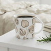 A white ceramic mug with a beige botanical leaf pattern sits on a white table in a bright bedroom setting, with a bed, glasses on books, and a sprig of greenery in the background.