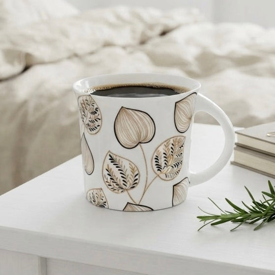 A white ceramic mug with a beige botanical leaf pattern sits on a white table in a bright bedroom setting, with a bed, glasses on books, and a sprig of greenery in the background.