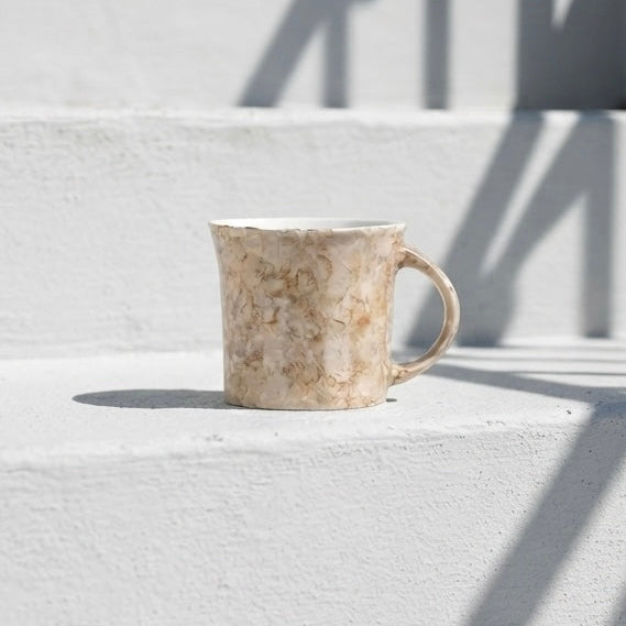 A small ceramic mug with a textured beige and brown marbled pattern sits on a white concrete staircase under bright sunlight, casting a shadow on the steps.