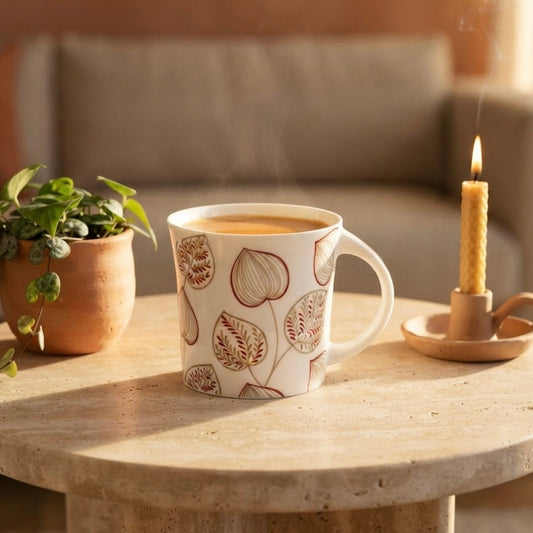 A white ceramic mug with a brown botanical leaf pattern sits on a round stone table in a cozy living room setting, next to a lit candle and a small potted plant.
