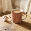 A pink ceramic mug with brown polka dots sits on a rustic wooden table in a cozy, softly lit living room setting, next to a small bowl of stones and cotton bolls.