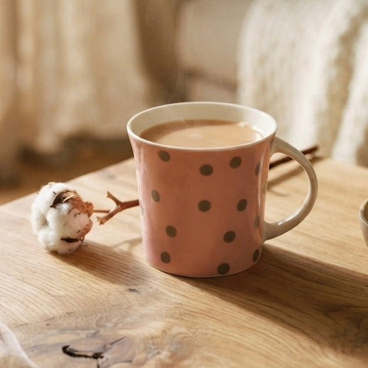 A pink ceramic mug with brown polka dots sits on a rustic wooden table in a cozy, softly lit living room setting, next to a small bowl of stones and cotton bolls.