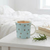 A light blue ceramic mug with brown polka dots sits on a white bedside table next to a stack of books and a rosemary sprig, with a softly blurred white bed and window in the background.