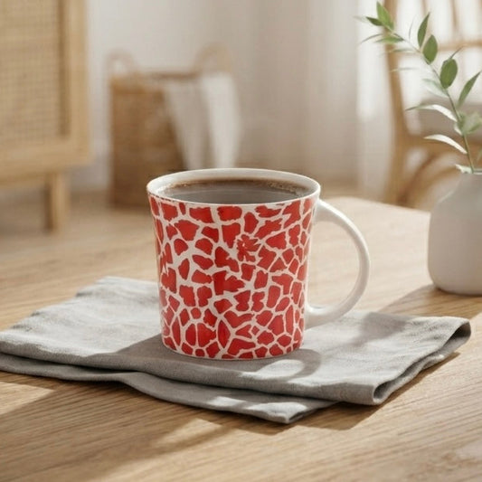 A ceramic mug with a red and white cracked-glass pattern sits on a folded gray napkin on a light wood table in a bright, modern living room setting.