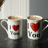 Two white ceramic mugs filled with coffee sit on a dark tabletop. Each mug is printed with the phrase 'I' followed by a large red heart and 'You' in black text.