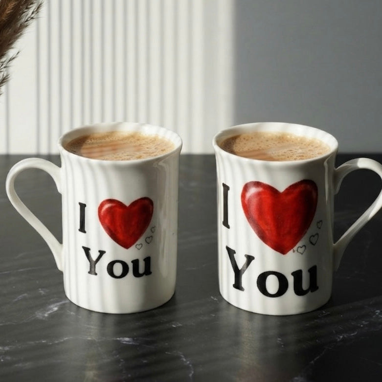 Two white ceramic mugs filled with coffee sit on a dark tabletop. Each mug is printed with the phrase 'I' followed by a large red heart and 'You' in black text.