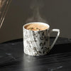 A white ceramic mug with a silver line-art pattern, filled with a steaming coffee or latte, set on a dark marble surface next to dried pampas grass.