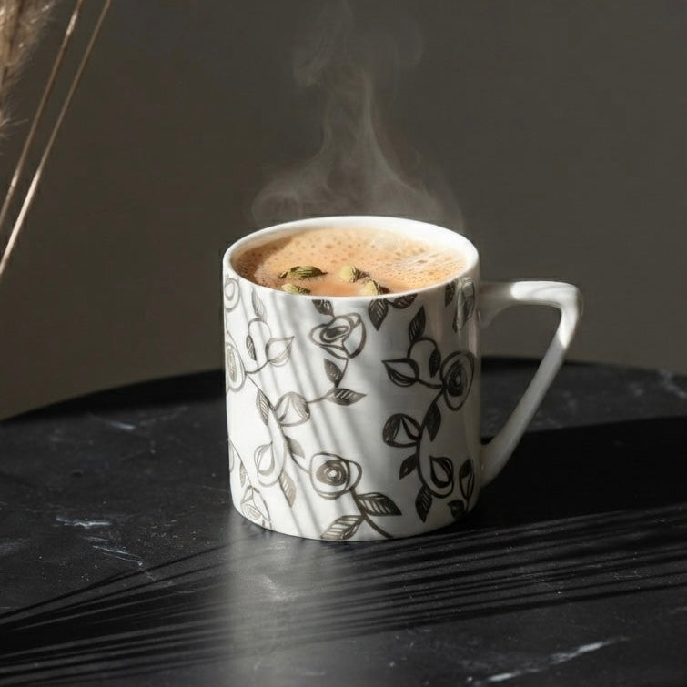 A white ceramic mug with a silver line-art pattern, filled with a steaming coffee or latte, set on a dark marble surface next to dried pampas grass.