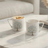 Two white ceramic mugs with blue and gray tree illustrations sit on a white marble table in a bright, modern living room setting. The scene includes a sofa in the background, a small glass vase with pampas grass, a lit candle in a gold holder, and a stack of books.