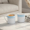 Two ceramic mugs with light blue and white gingham and polka dot patterns sit on a white marble coffee table in a bright, modern living room setting.