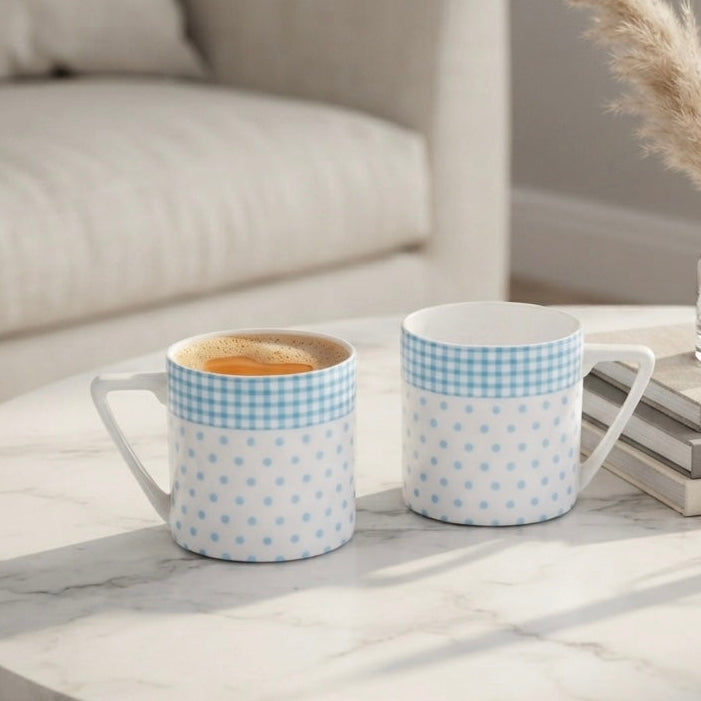 Two ceramic mugs with light blue and white gingham and polka dot patterns sit on a white marble coffee table in a bright, modern living room setting.