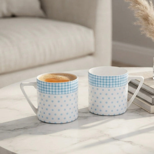 Two ceramic mugs with light blue and white gingham and polka dot patterns sit on a white marble coffee table in a bright, modern living room setting.