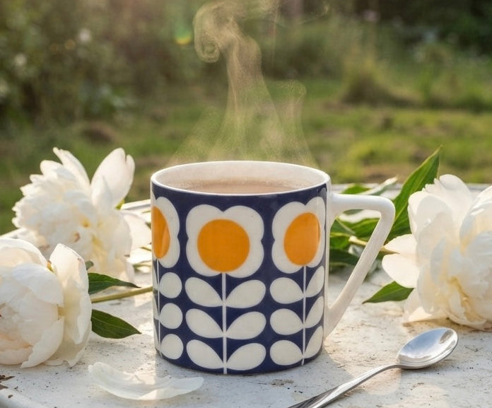 A ceramic mug with a navy blue and white floral pattern and yellow flower centers sits on a weathered white table outdoors, with white flowers and a spoon around it and steam rising in a sunlit garden setting.