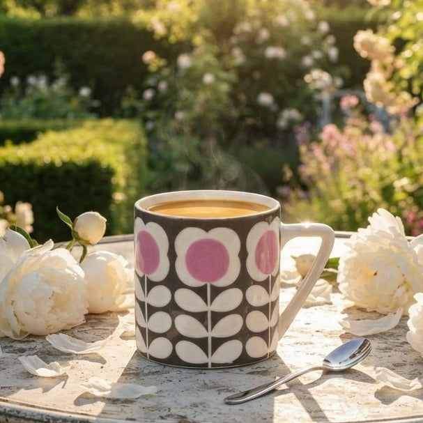 A patterned ceramic mug filled with a golden beverage sits on a distressed white metal garden table, surrounded by white flowers in a sunlit garden setting.