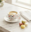 A white ceramic coffee cup and saucer with a black, red, and orange vertical dot pattern, filled with coffee, set on a marble countertop next to macarons and a folded napkin.