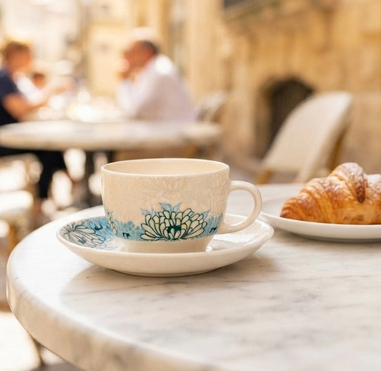 A white ceramic coffee cup with a blue floral pattern sits on a matching saucer on a round marble table, next to a croissant on a small plate, with a blurred outdoor cafe scene in the background.