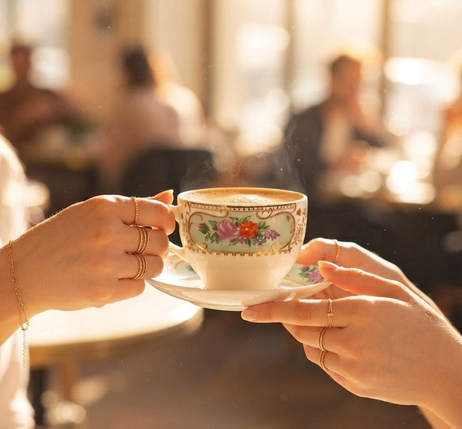 A close-up lifestyle shot of two hands holding a vintage-style floral gold teacup and saucer in a warm, sunlit cafe setting.