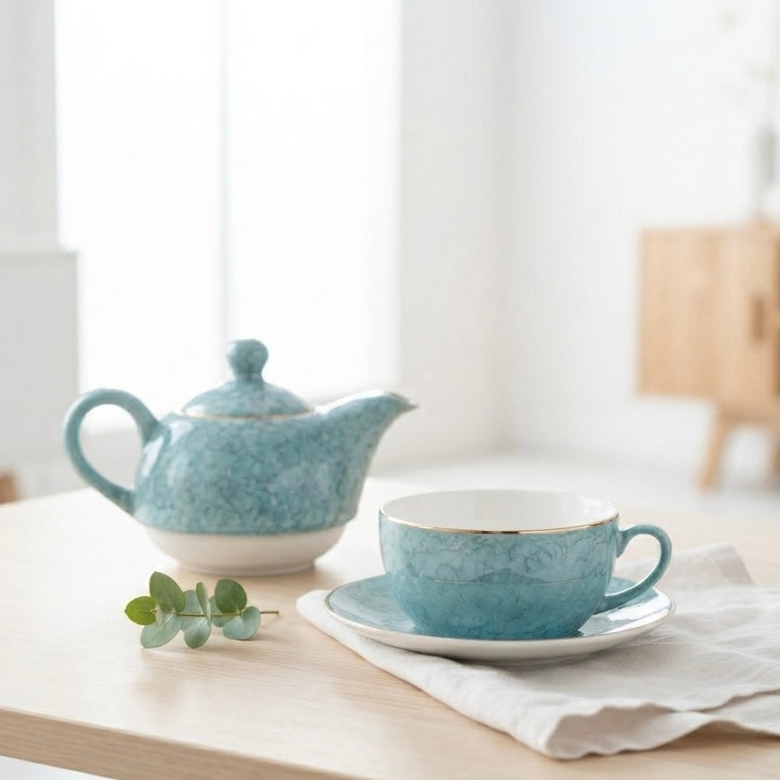 A light blue speckled ceramic teapot and matching cup with saucer on a light wood table, styled with a white napkin and a small green sprig in a bright, modern room.