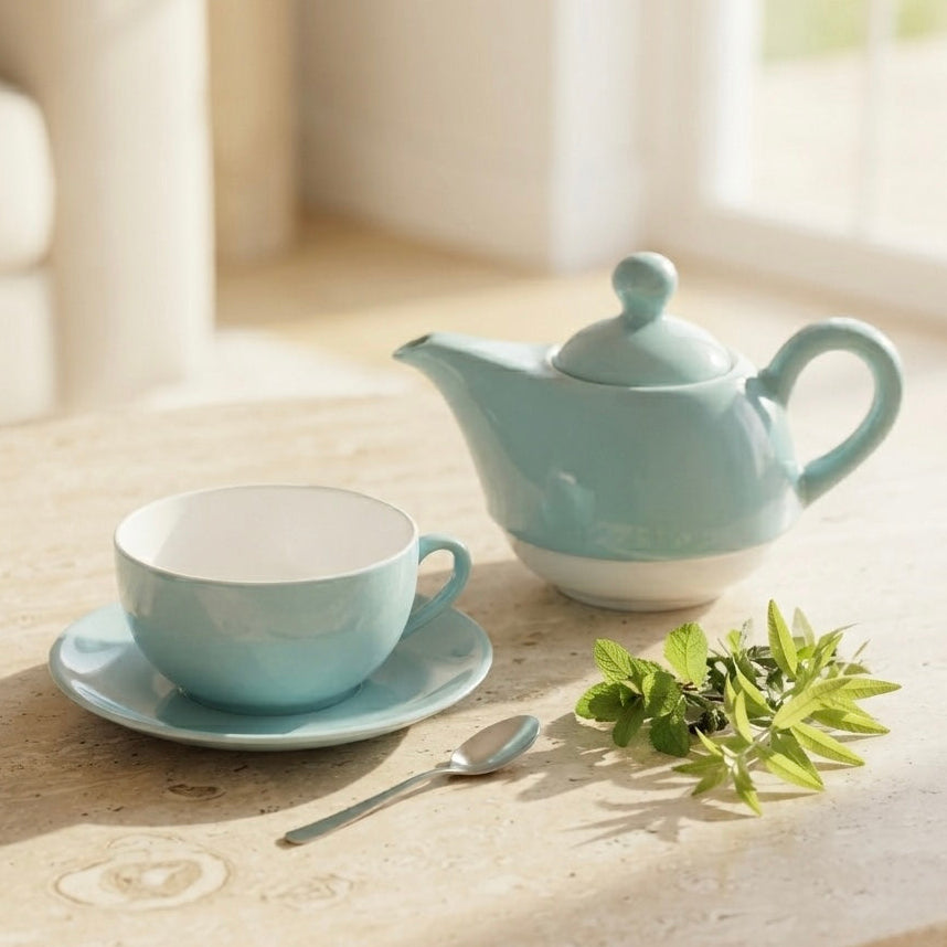 A light blue ceramic teapot with a matching cup and saucer, styled on a round stone table with a spoon and green leaves in a bright, softly blurred interior setting.