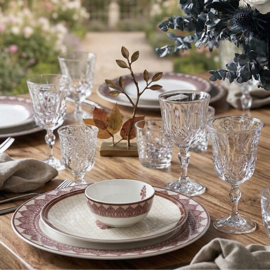 Dining table set with patterned plates, glasses, and a floral arrangement outdoors.